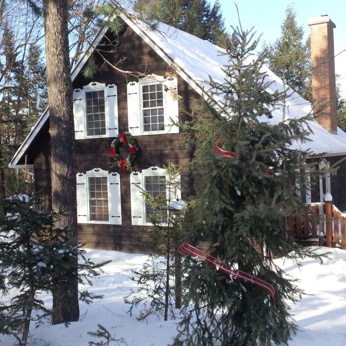 Snow-covered A-frame cabin with 4 white-shuttered windows in the woods with a Christmas wreath on the cabin
