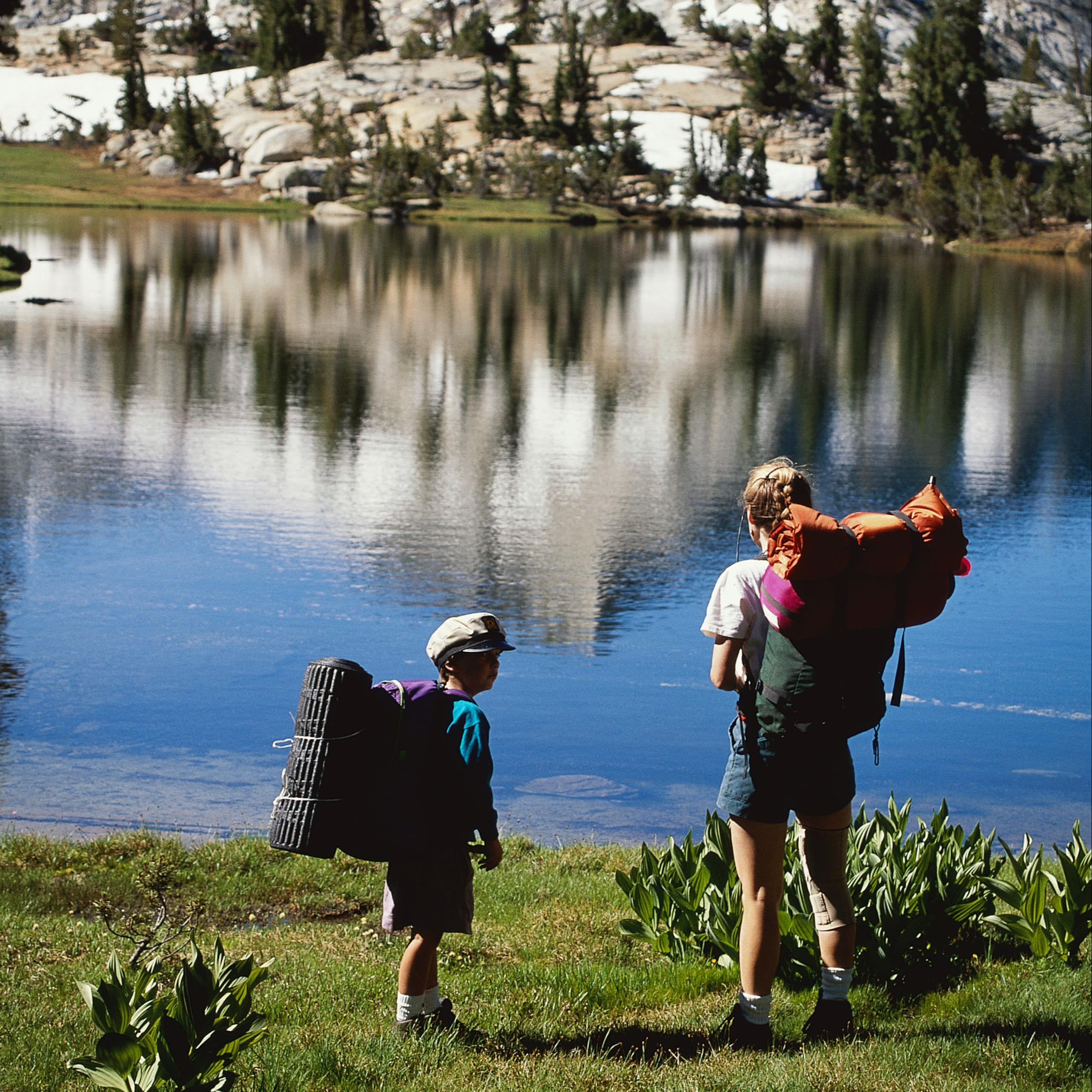 Mom and son hikers stop to take in the scenery of a beautiful lake with a snow covered mountain behind it. 