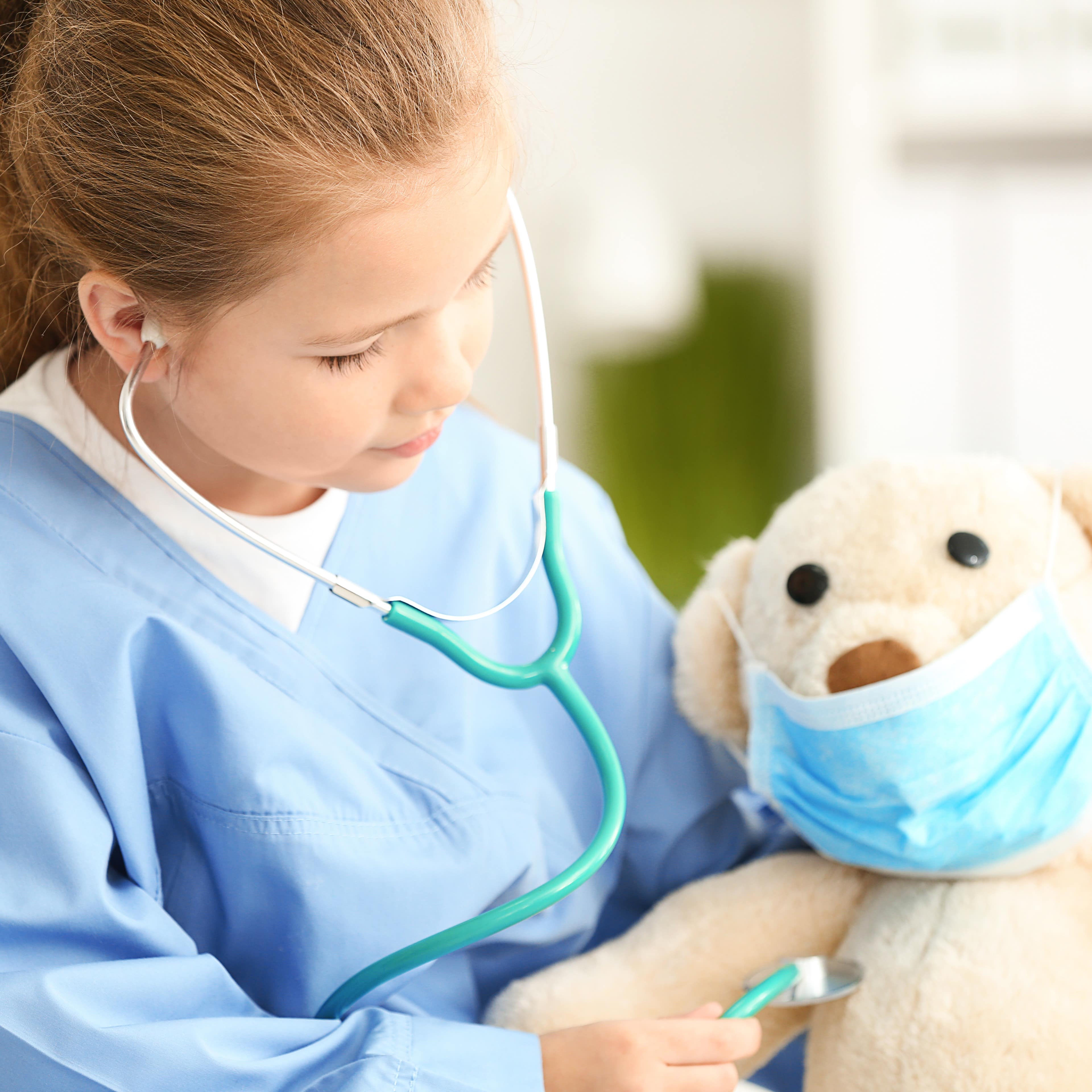Cute little girl in doctor uniform playing with toy bear in hospital