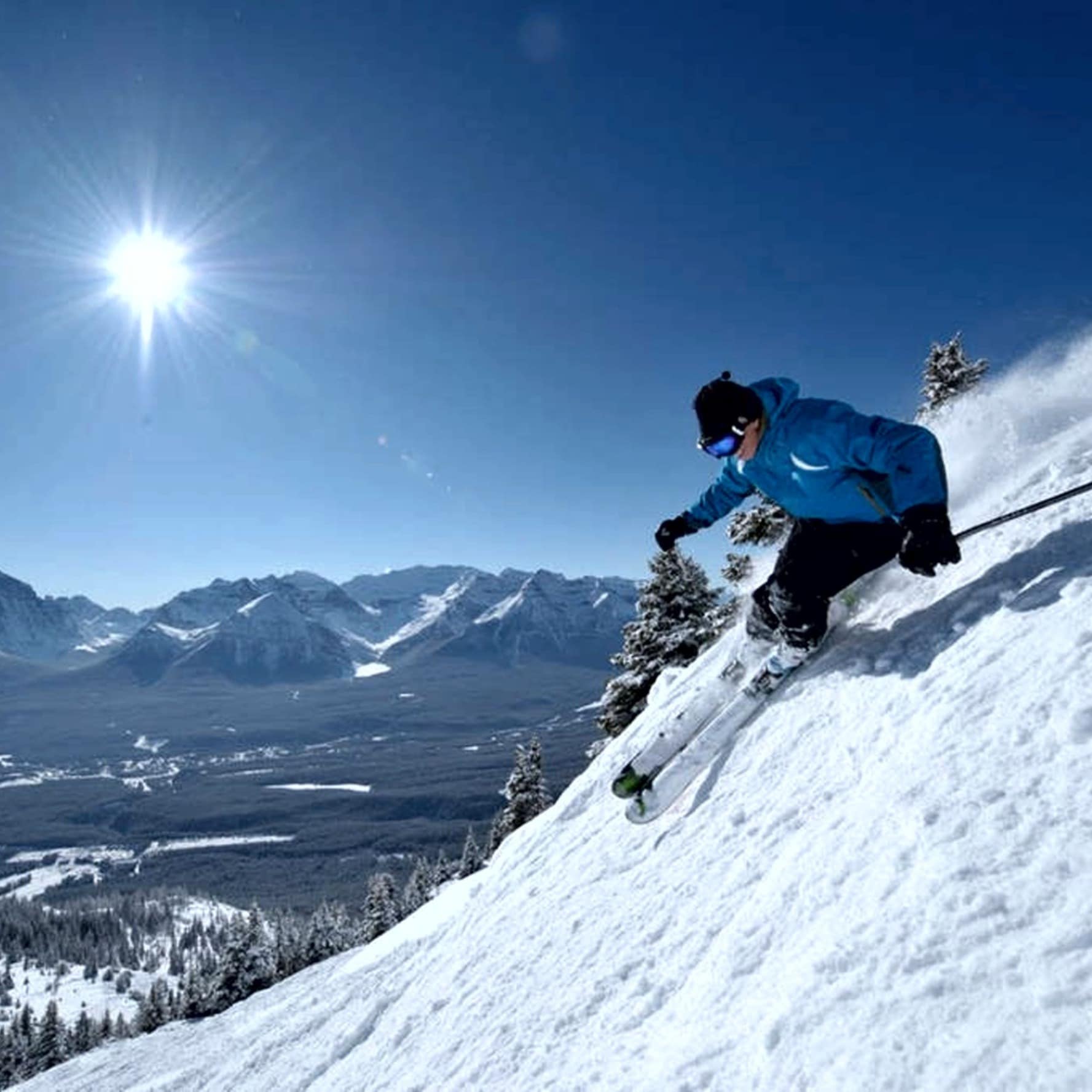 Skier coming down a steep ski slope on a sunny day with mountains in the background.