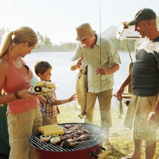 Family gathered around a grill with the lake behind them, while their son inspects a fish that was just caught in the lake.