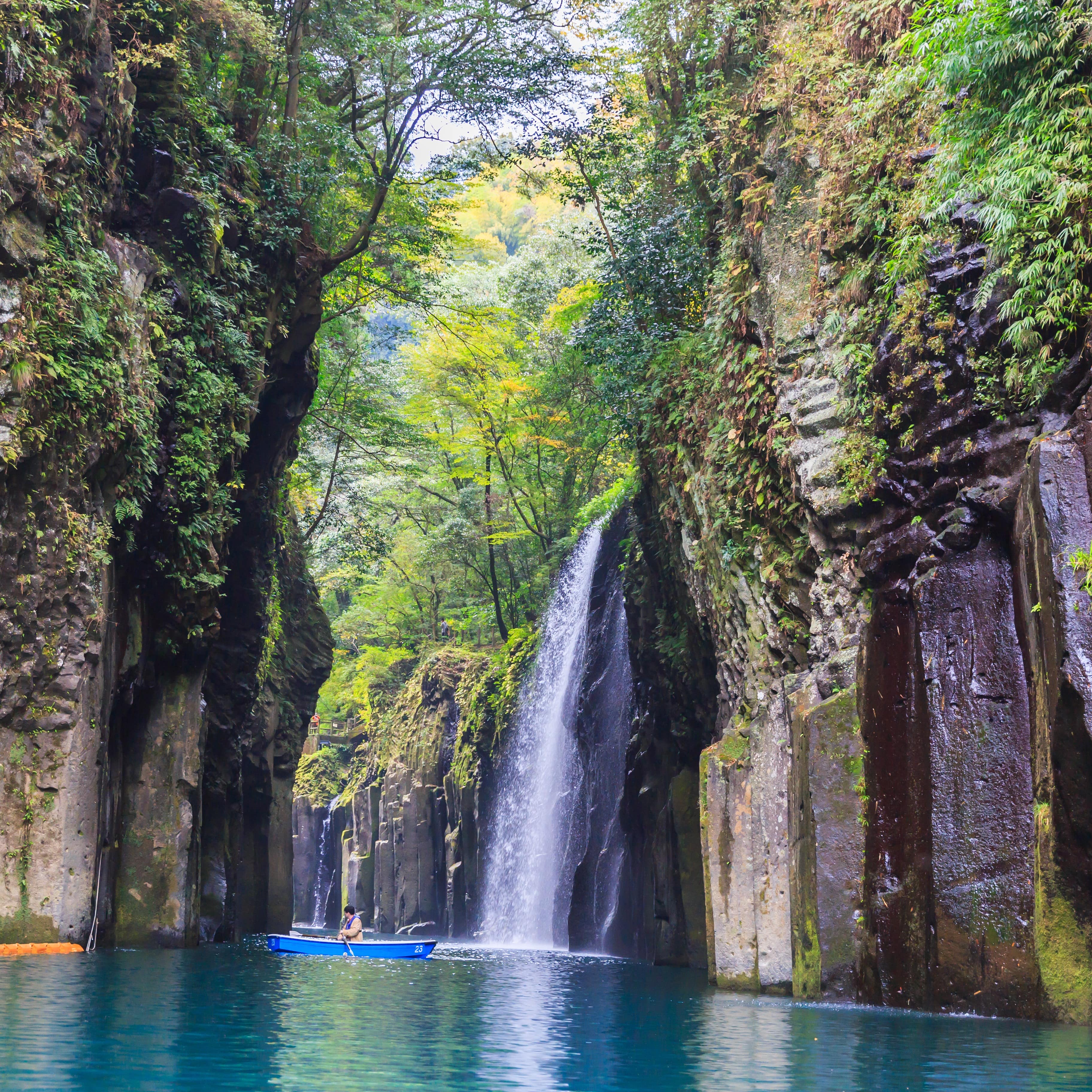 Couple in a rowboat taking in the scenery of a waterfall cascading down a cliff with beautiful green foliage surrounding them from above.