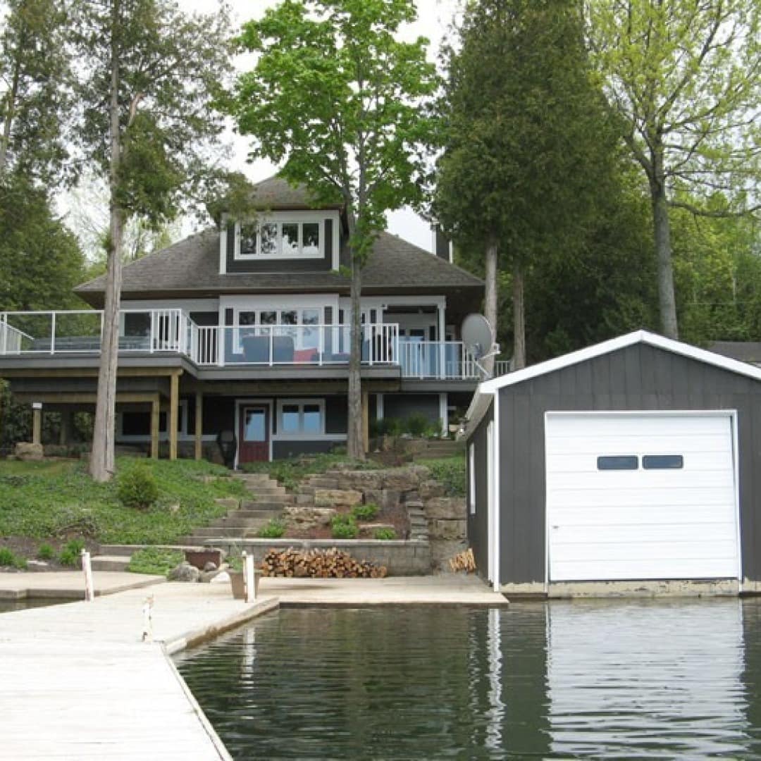 View from dock on lake of two story cottage with wrap around porch nestled in trees