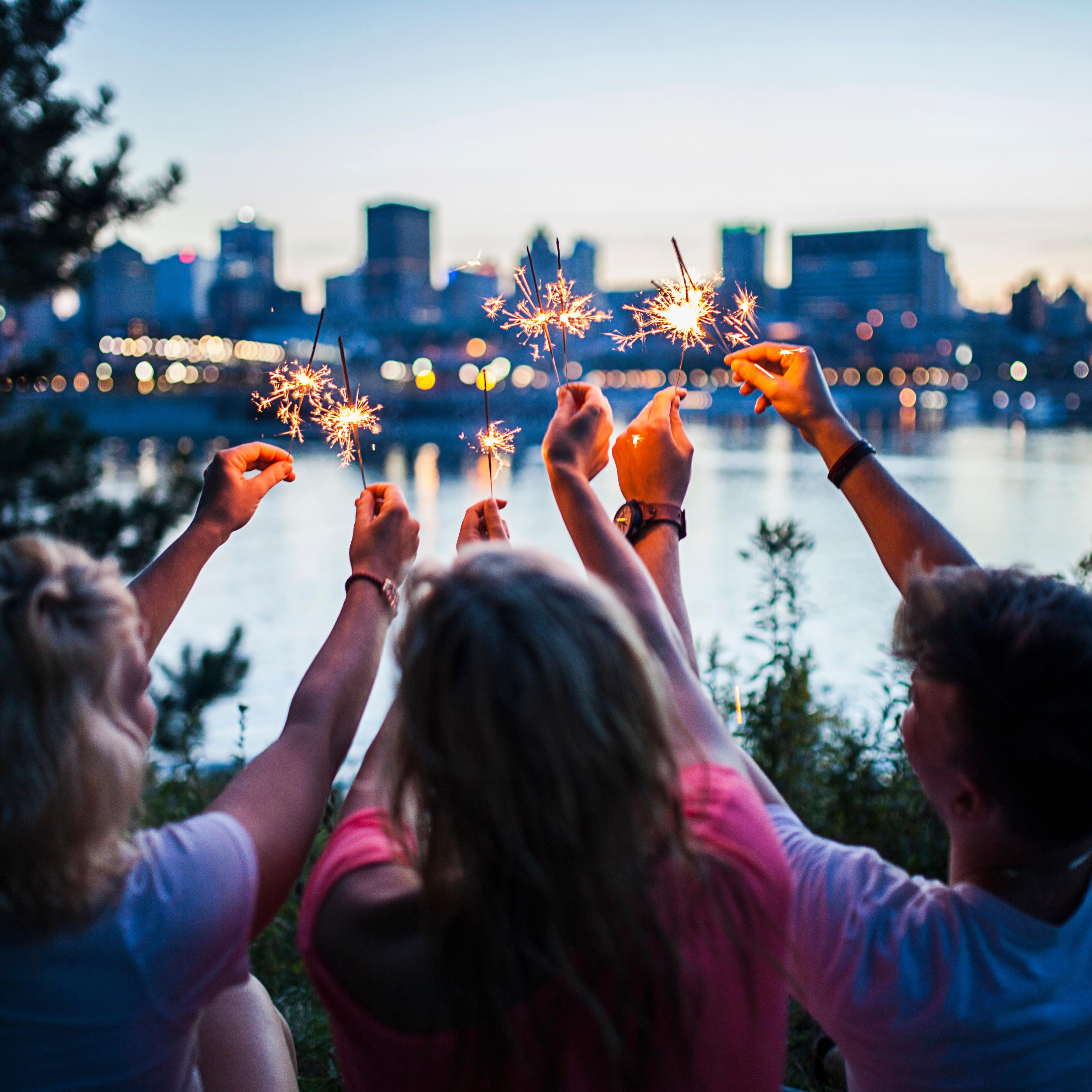 Three people holding sparklers together at dusk with a blurred city in the background