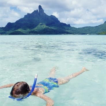 Child snorkeling in the clear water with a mountain in the background