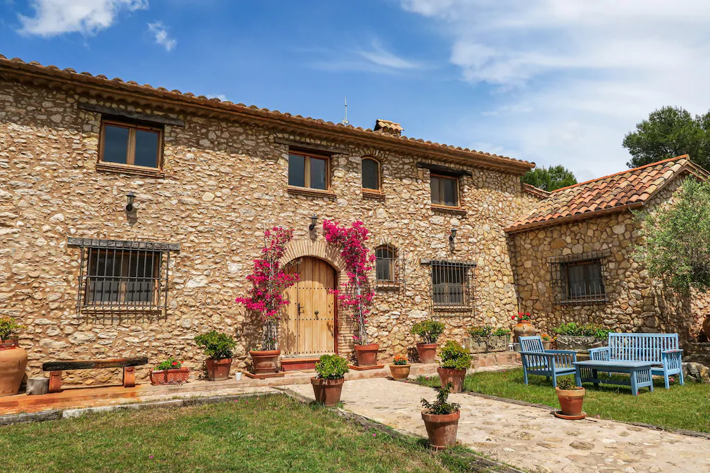 Entrada a una finca de piedra con zona ajardinada y bancos en tonos azules