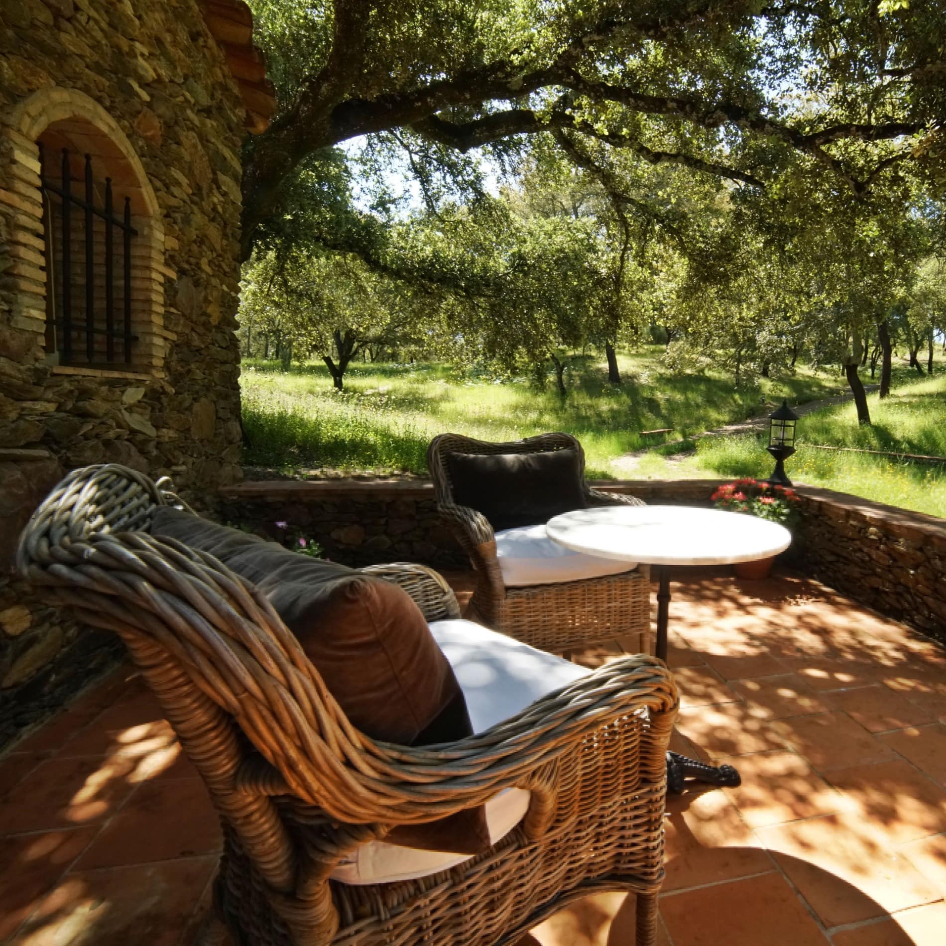 Porche de casa de piedra bajo un árbol, con suelo de barro, sillones de mimbre y una mesita