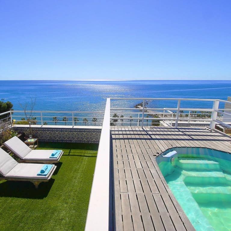 Dos tumbonas en una terraza con césped artificial, jacuzzi elevado y vistas al mar