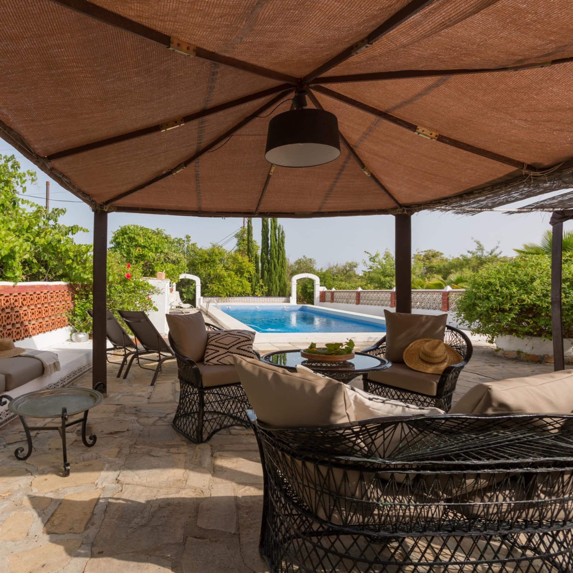 Zona exterior de una casa con suelo de piedra, muebles de mimbre, plantas y piscina al fondo