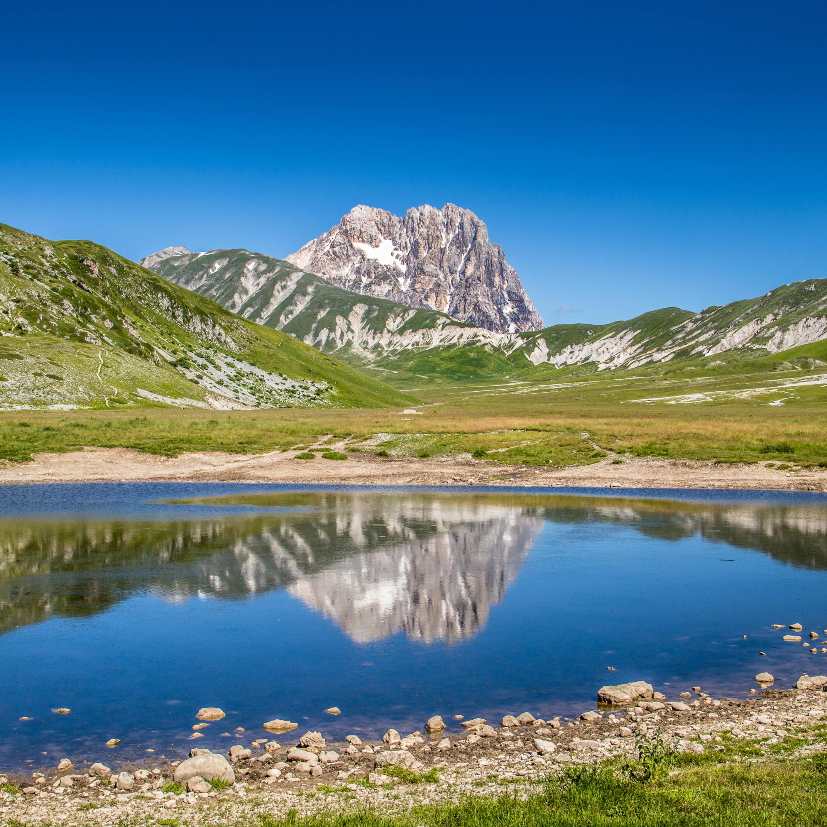 Il massiccio del Gran Sasso che svetta nel cielo terso riflesso in un lago di montagna dall’acqua cristallina