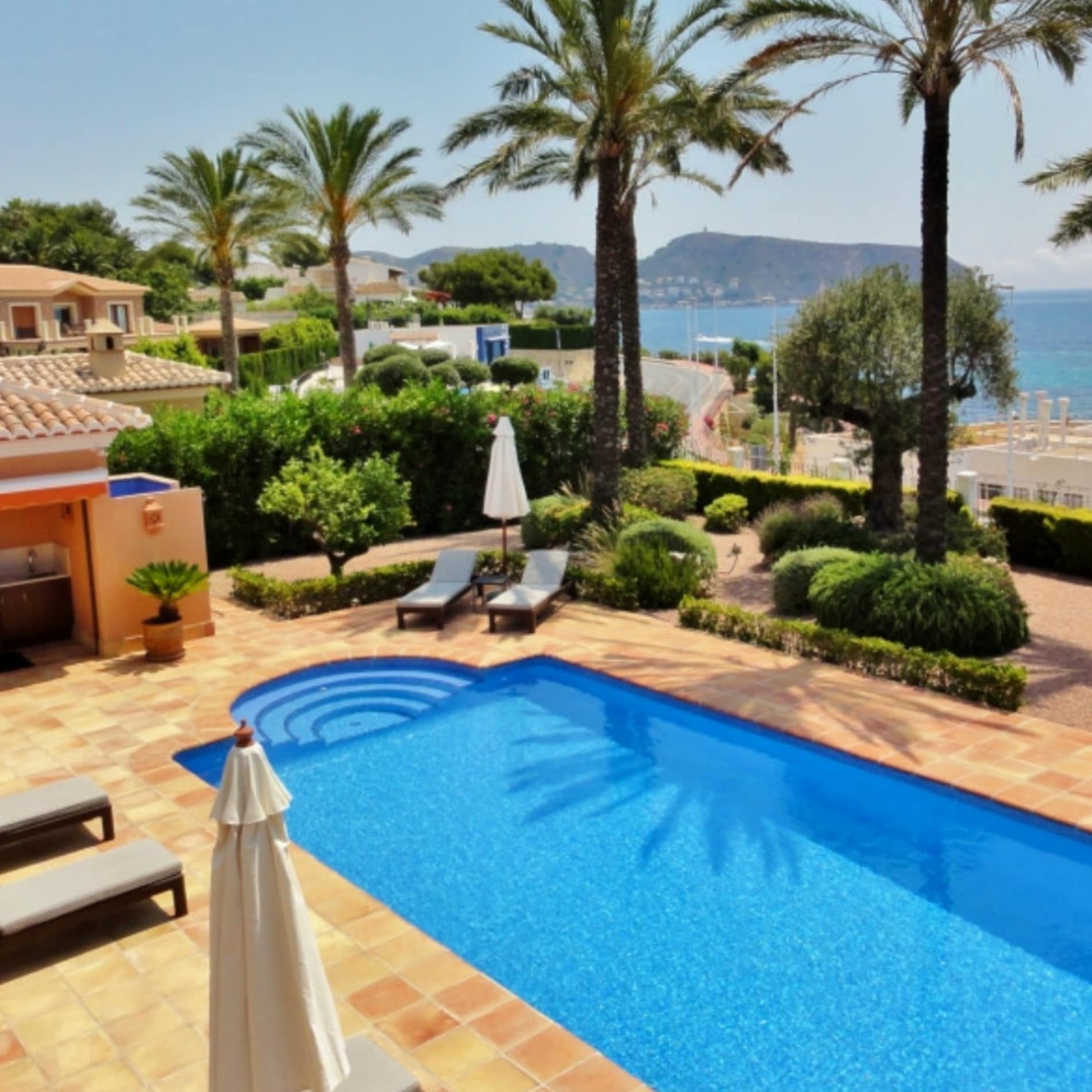 Zwembad met helder blauw water omgeven door zonnig terras met ligbedden', palmbomen en zeezicht