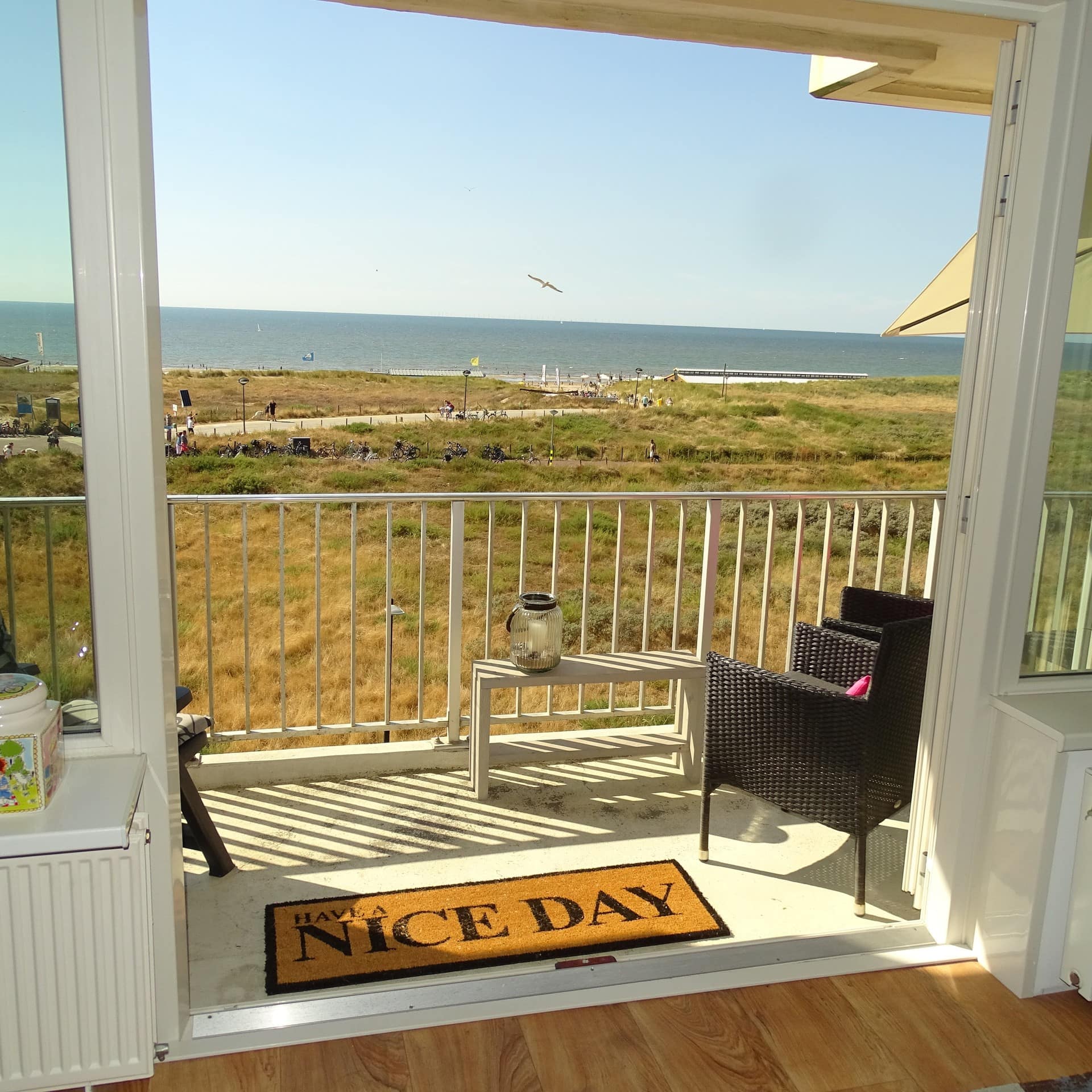 Uitzicht op de duinen en de zee vanuit de kamer van een vakantiehuisje. Op de voorgrond openslaande deuren naar een balkon