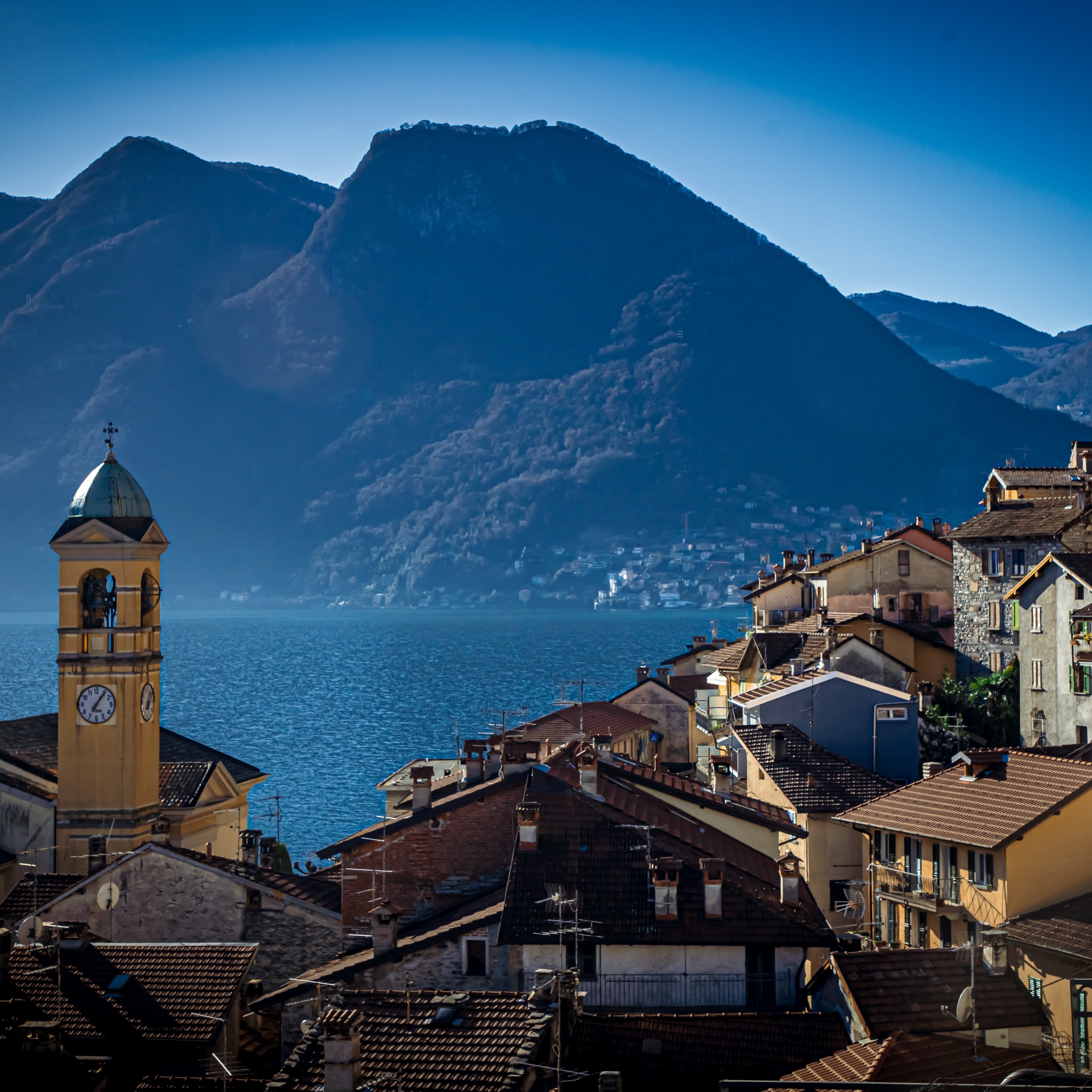 Een doorkijk naar het Comomeer vanuit een rustiek Italiaans stadje gelegen op een heuvel, met op de achtergrond heuvelachtig landschap. 