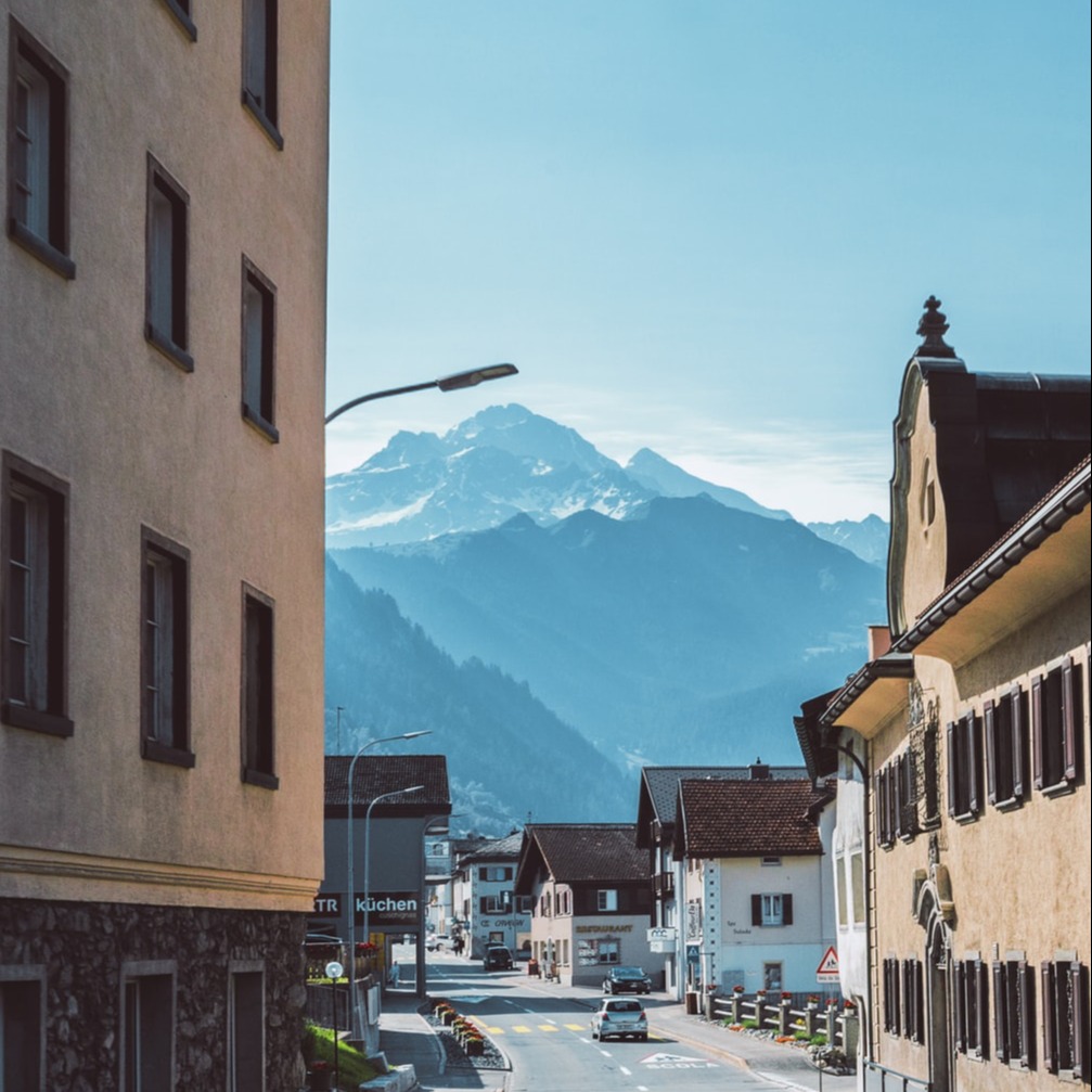 Een rustig dorpsstraatje met een weg er doorheen met bergen op de achtergrond onder een blauwe hemel