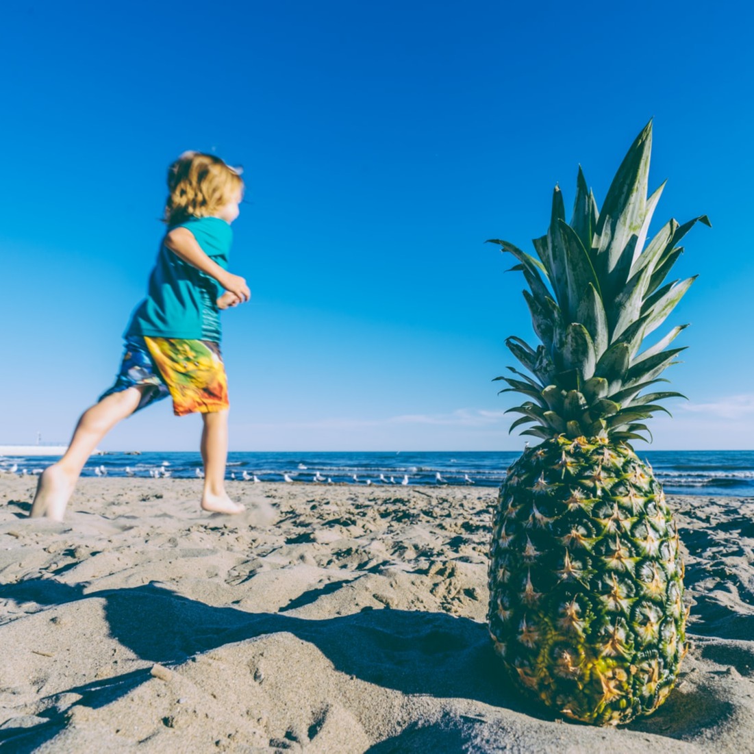 Een rennend kind in een korte broek aan het strand met een ananas op de voorgrond in het zand 