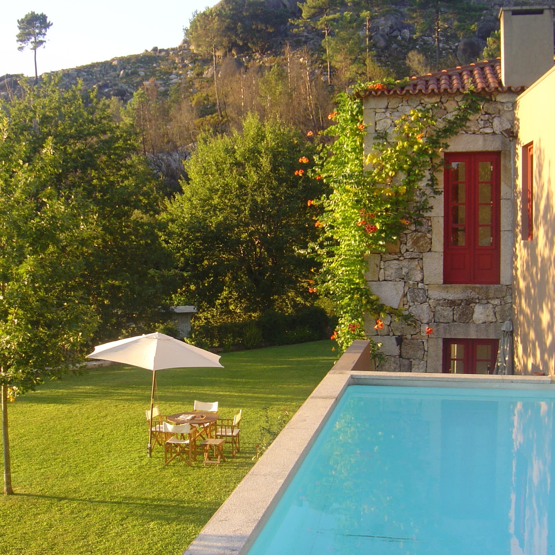 Piscina, relvado, árvores, mesa e cadeiras de madeira com guarda-sol; casa rústica em pedra com vista para o monte.