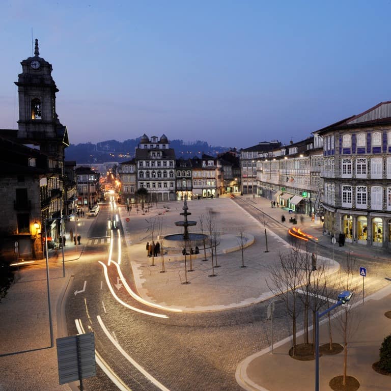 Centro histórico de Guimarães com edifícios antigos; à direita, parte de muralha com a inscrição “Aqui nasceu Portugal”.