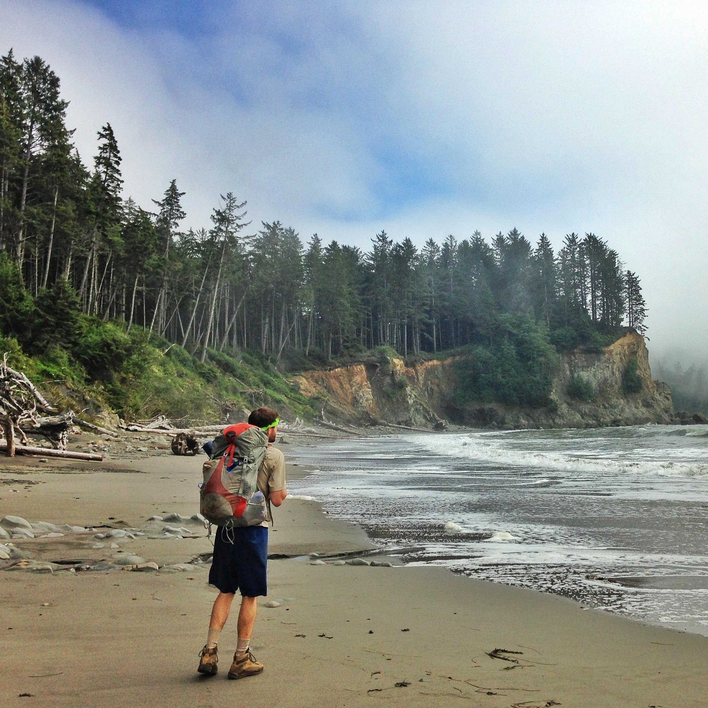 Man with backpack on windswept beach backed by pine trees