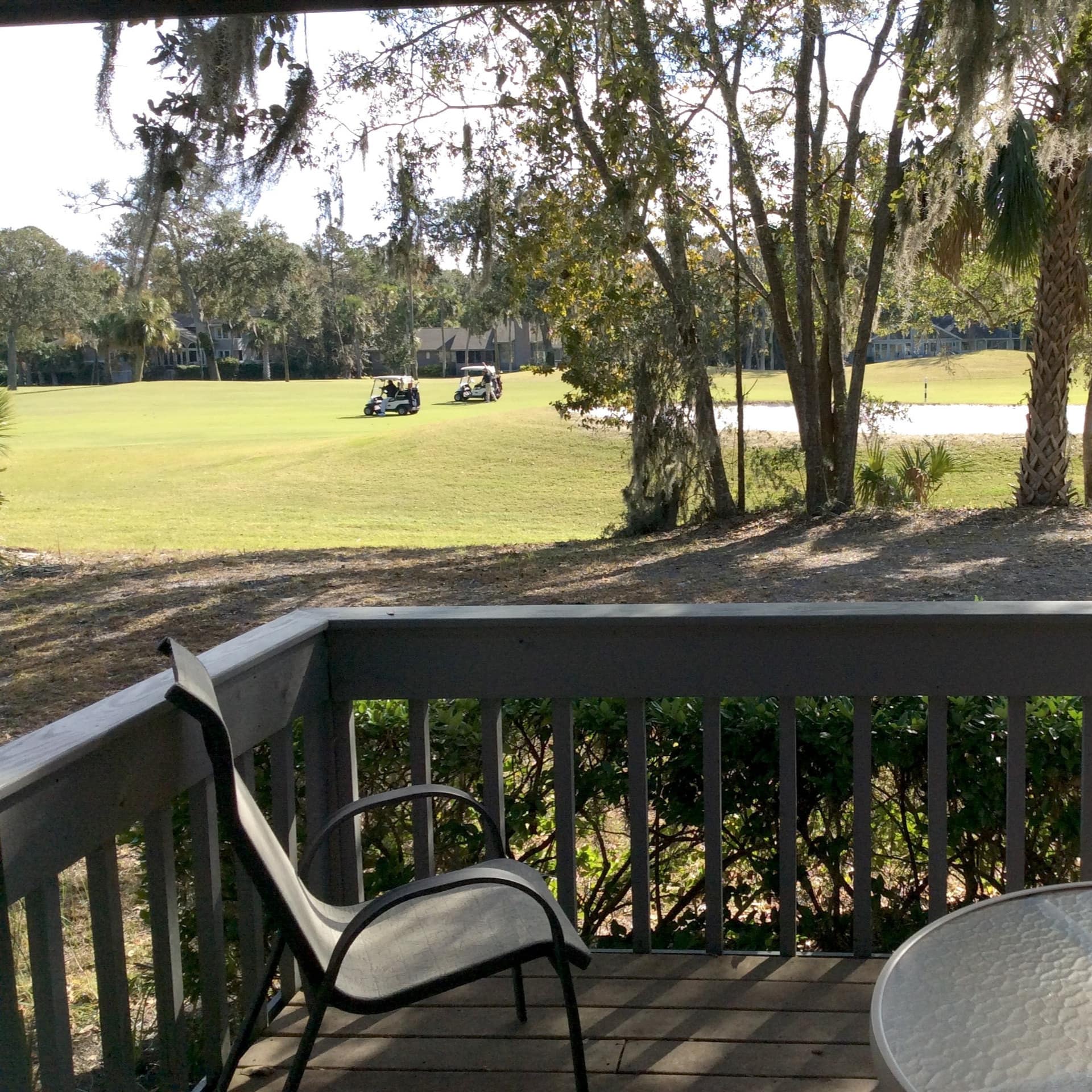 A furnished deck with a view of 2 golf carts driving across a tree-lined and sand-bunkered fairway.