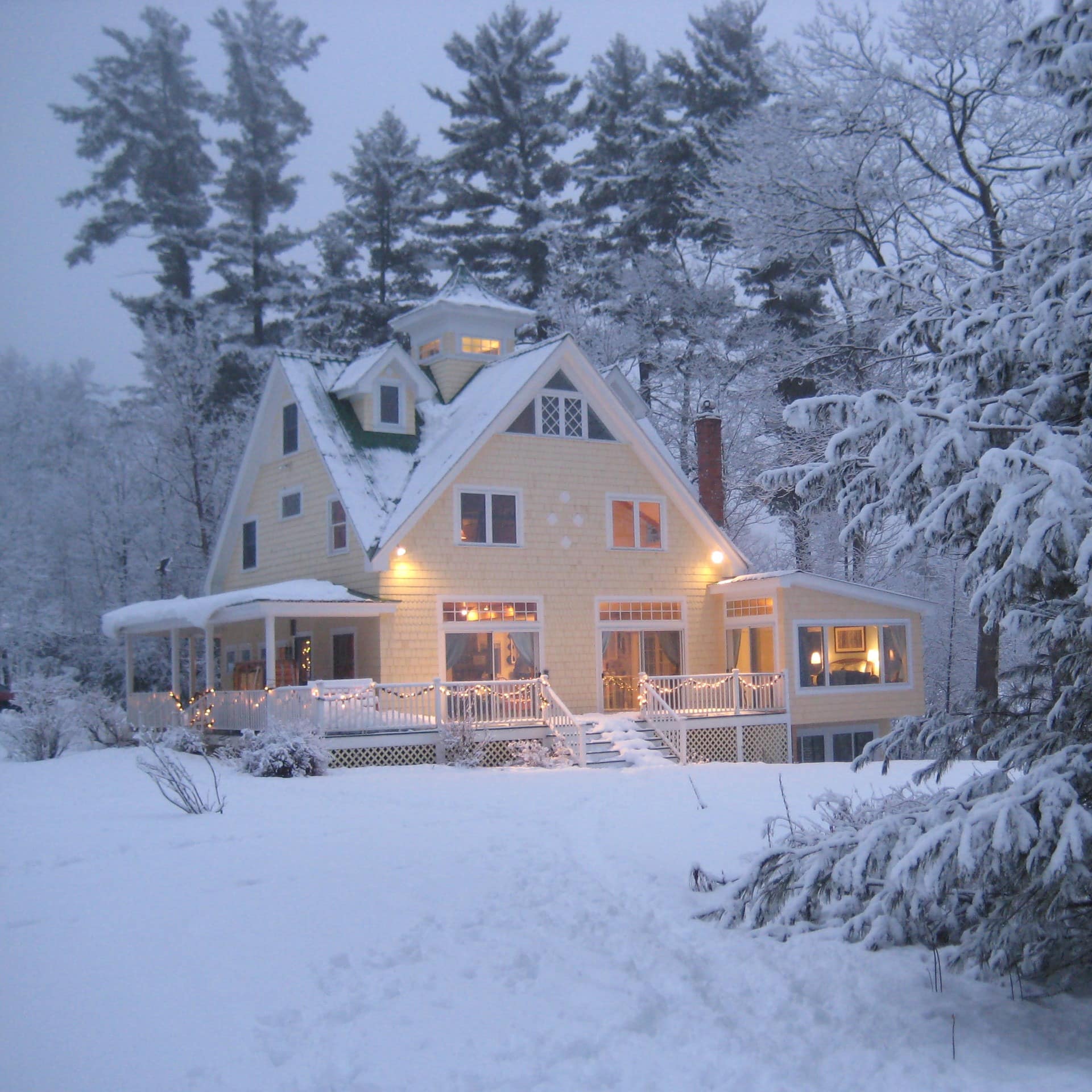 A yellow farmhouse rental in winter, surrounded by snow-covered trees and illuminated by Christmas lights