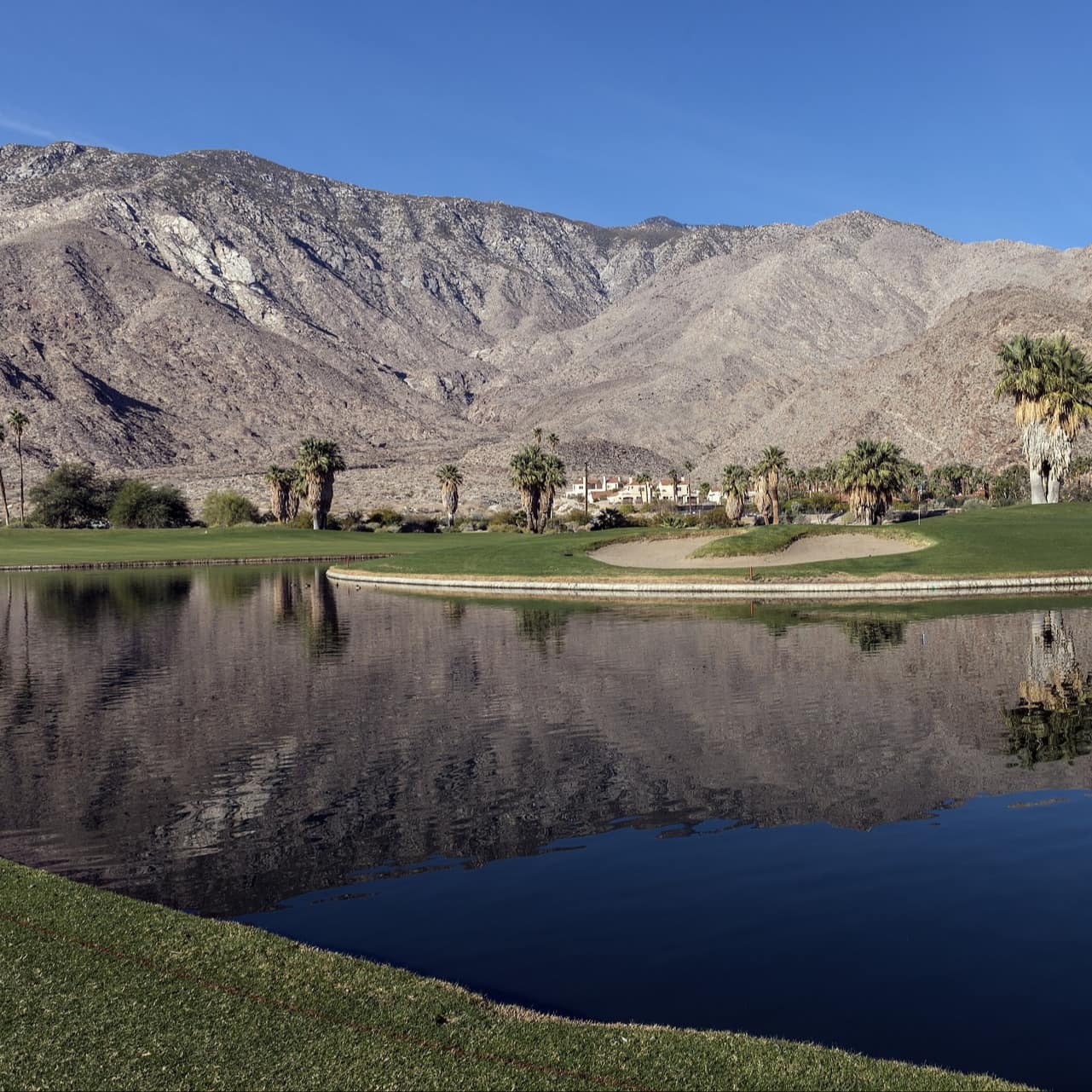 A body of water and a sand bunker in a golf course, with mountains in the background