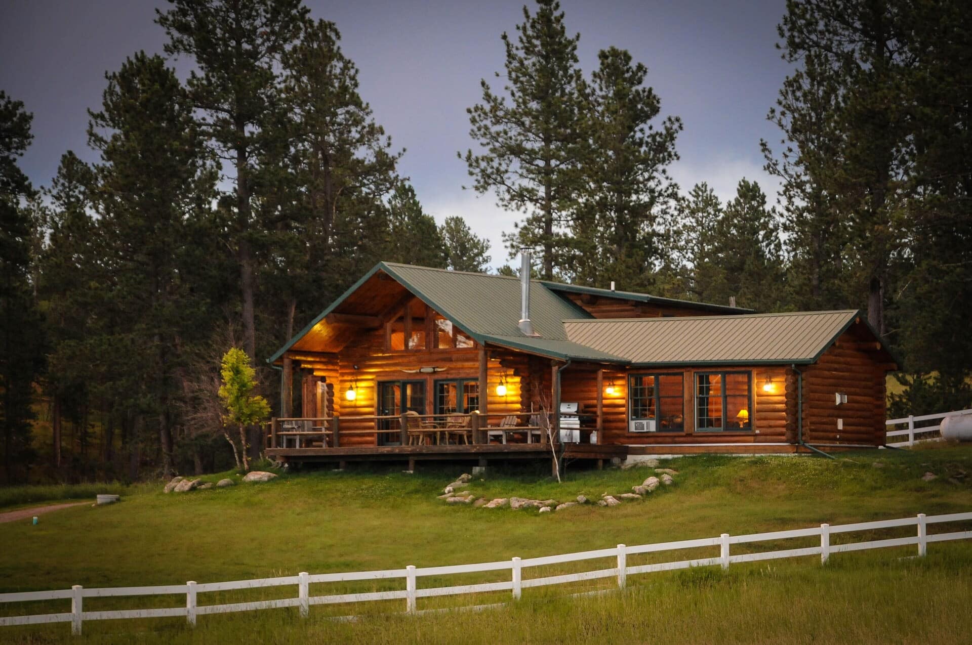 Log cabin nestled among tall trees with the porch lights on.