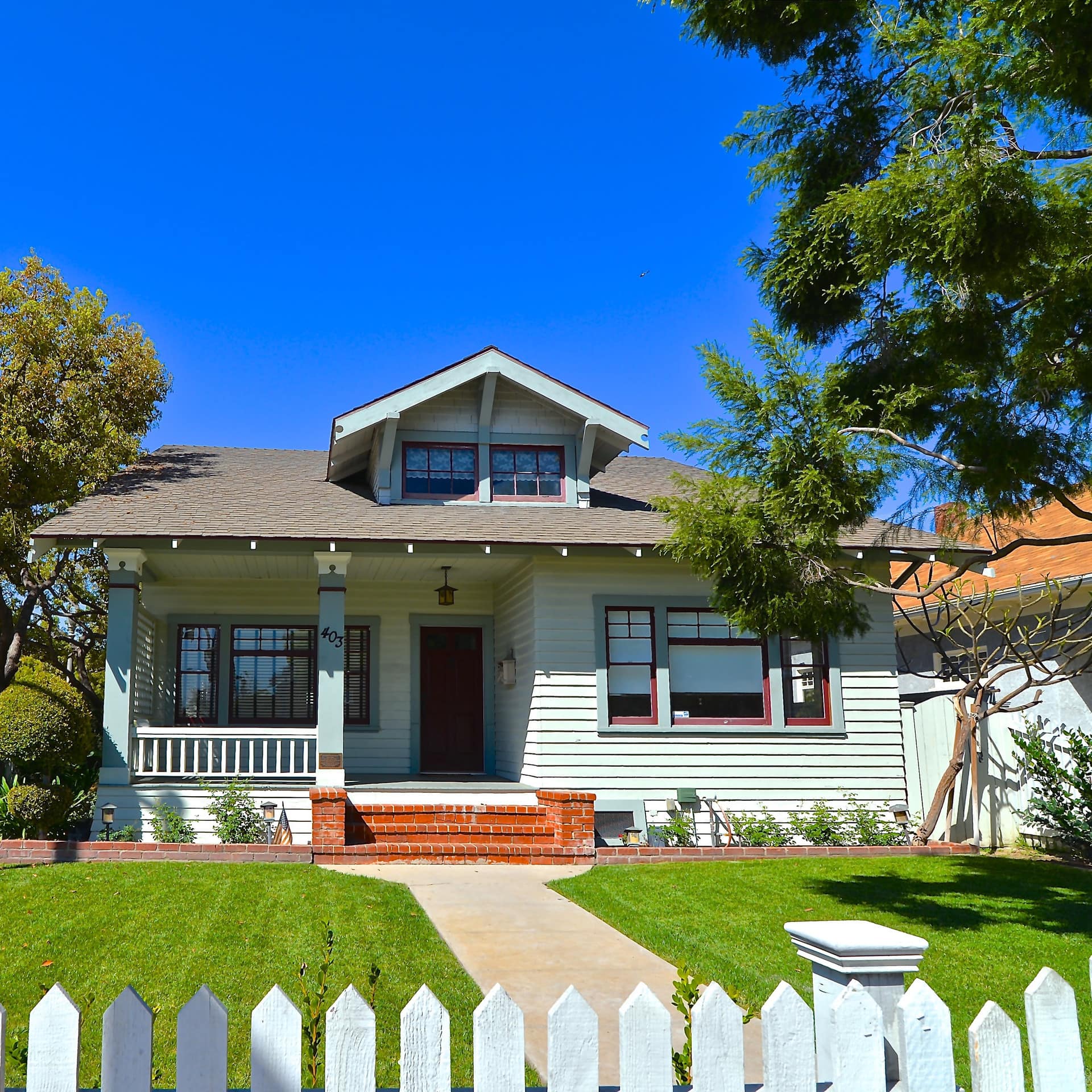 A quaint Craftsman bungalow on a manicured lawn surrounded by trees under a blue sky