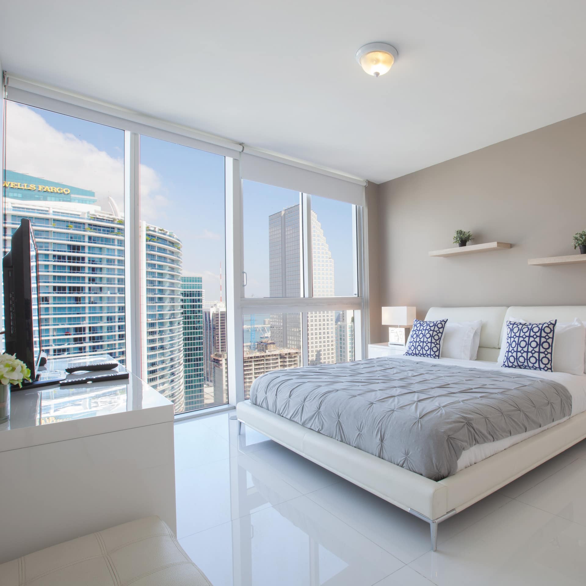 A furnished white bedroom with floor-to-ceiling windows providing views of skyscrapers