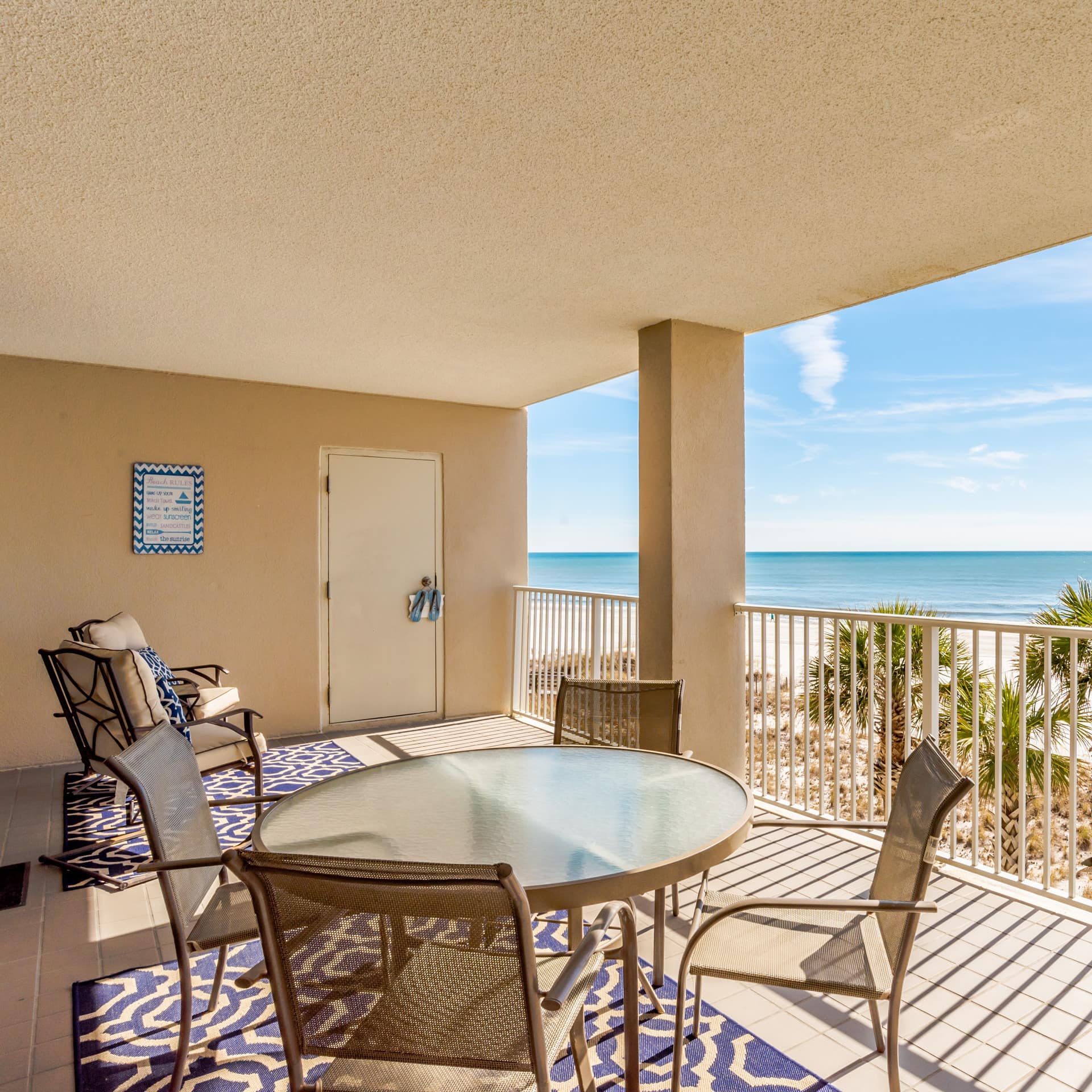 Alt text: A balcony with chairs and a table overlooks the glowing white sands and palm trees of Orange Beach