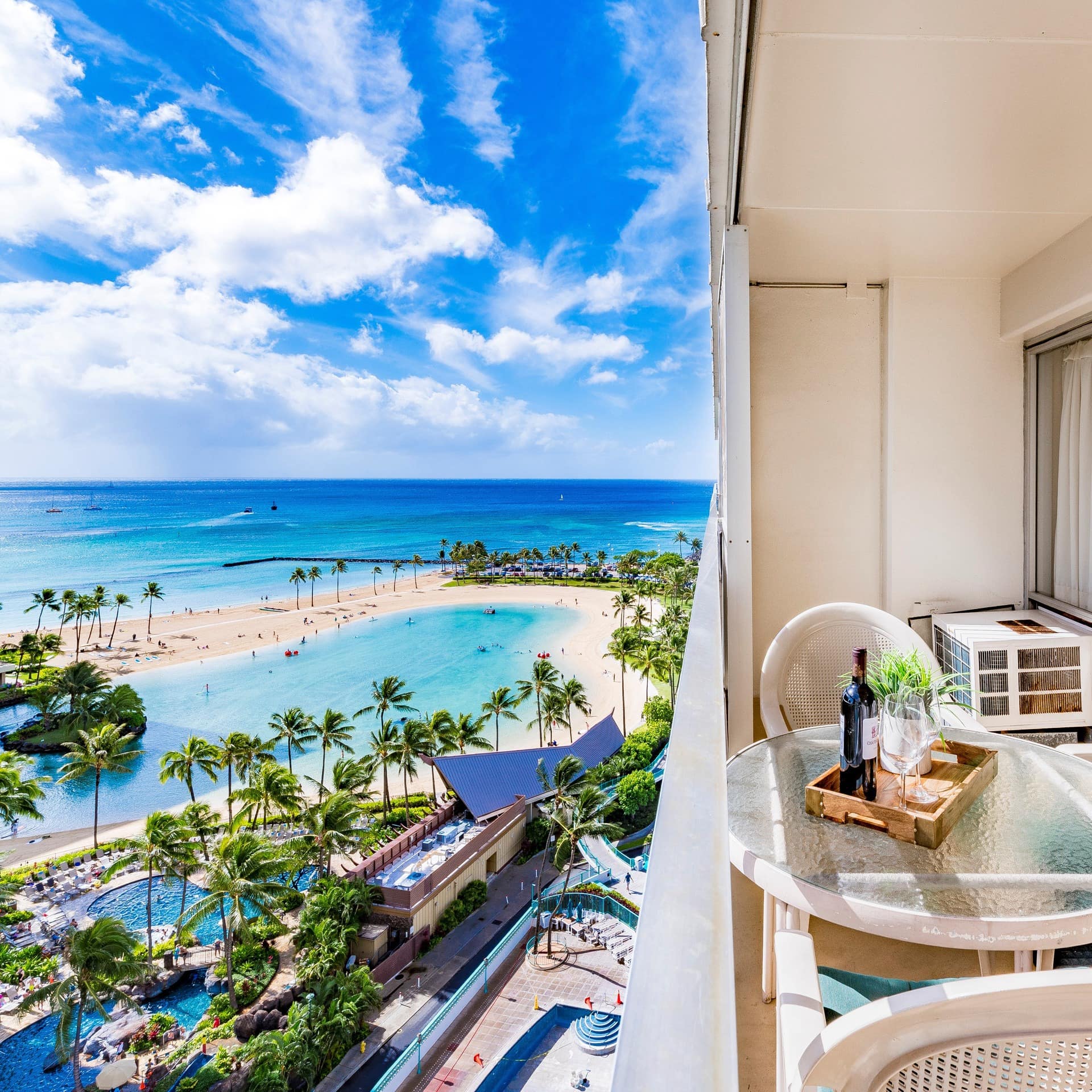 A furnished balcony overlooking pools and palm trees, with the blue ocean in the distance