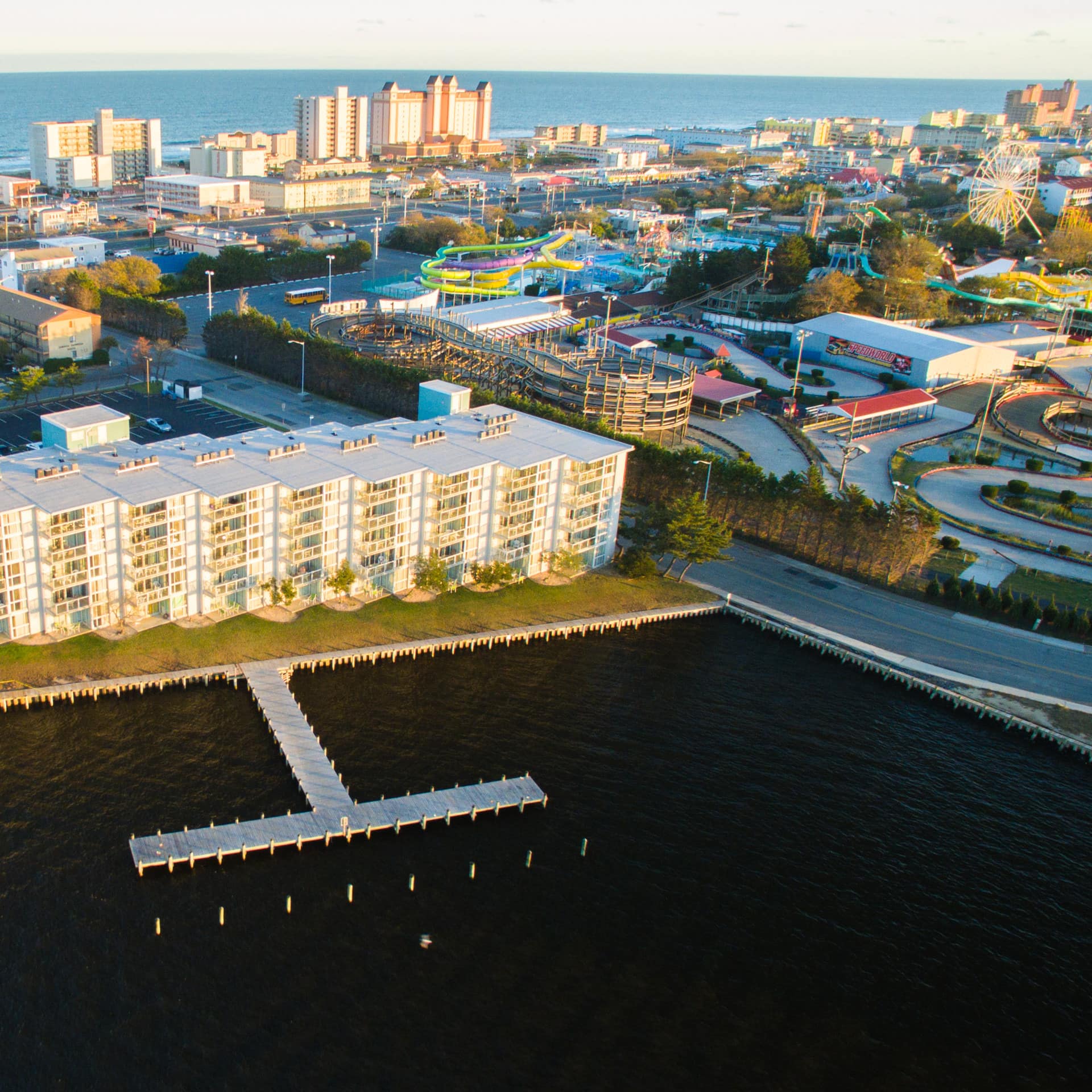 An aerial view of a condo building in the golden hour, with a dock on the foreground, plus an amusement park on the right side
