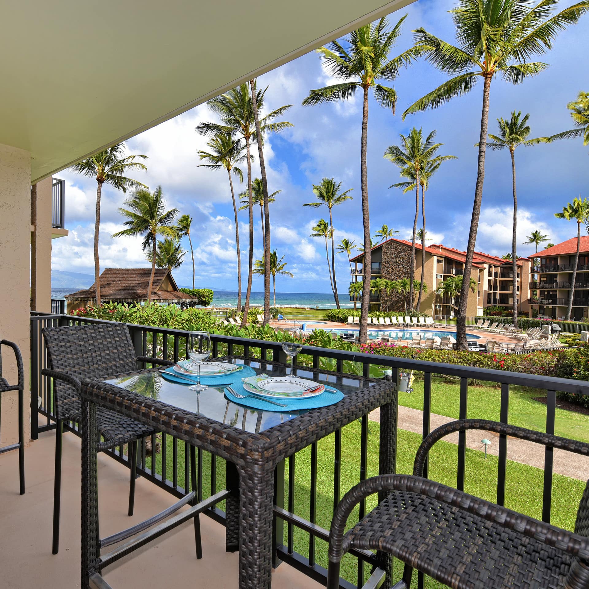 Table set for 2 on a deck that offers views of Papakea Resort’s outdoor pool, as well as partial ocean and mountain views