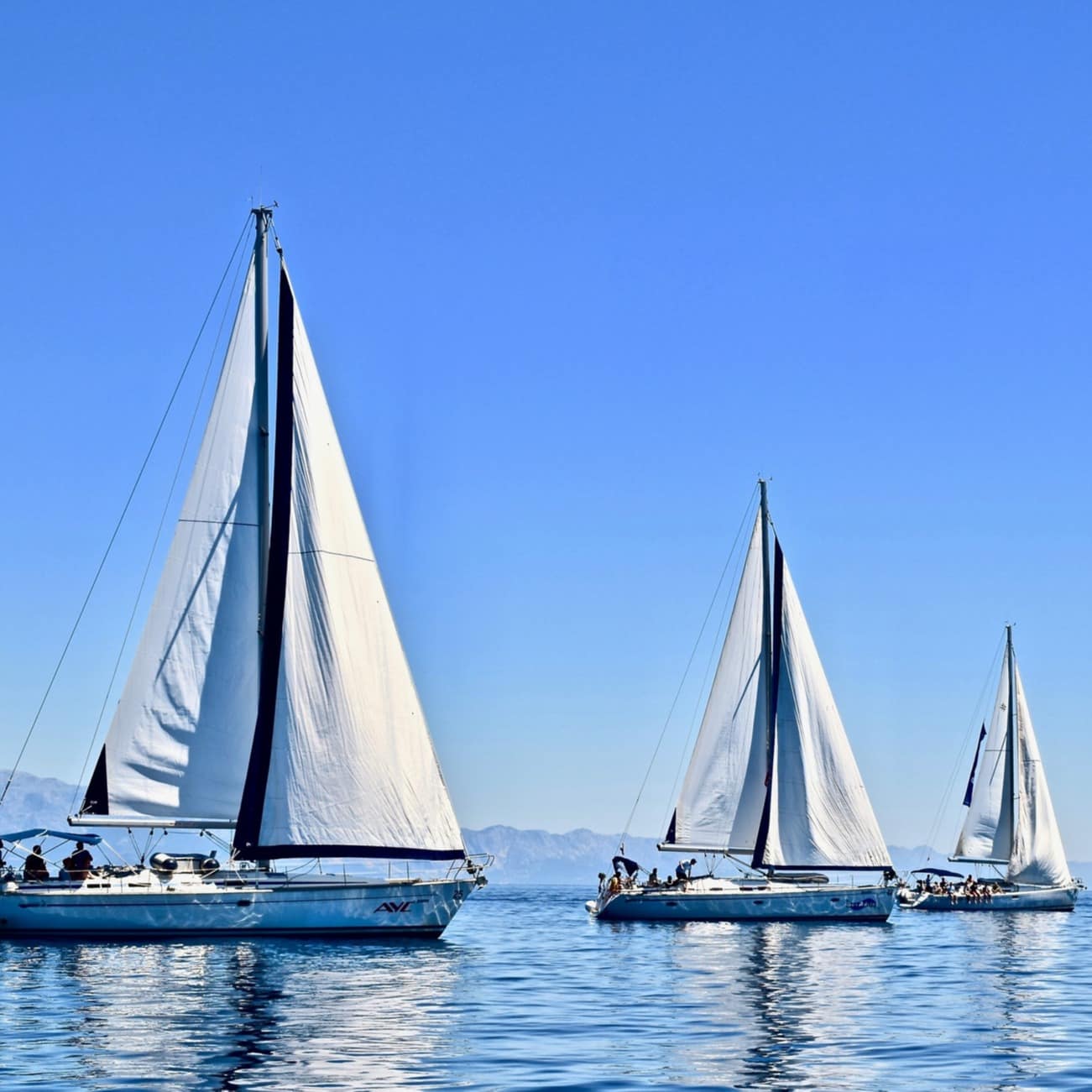 Three yachts in a row skim over an ocean that's framed by mountains