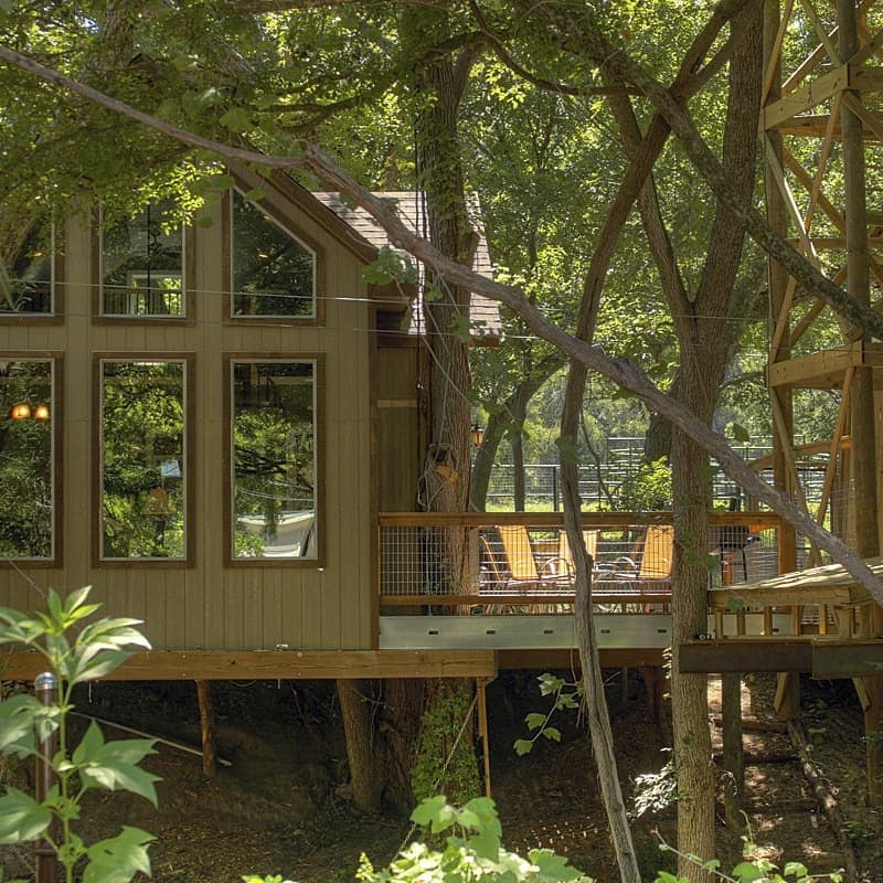 A treehouse with high windows and a deck with chairs sits in the woods of Texas