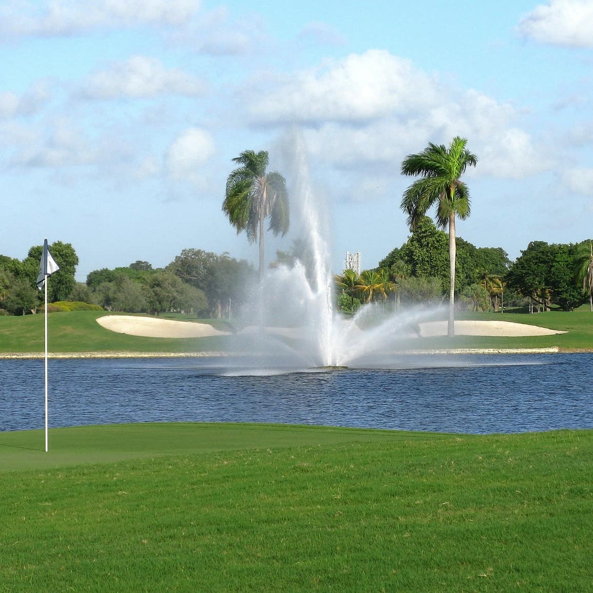 A fountain in the middle of a water trap in a golf course lined with palm trees