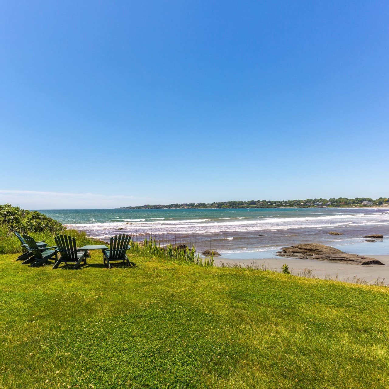A set of sunning chairs sit on a green lawn by a Rhode Island beach where the waves roll in