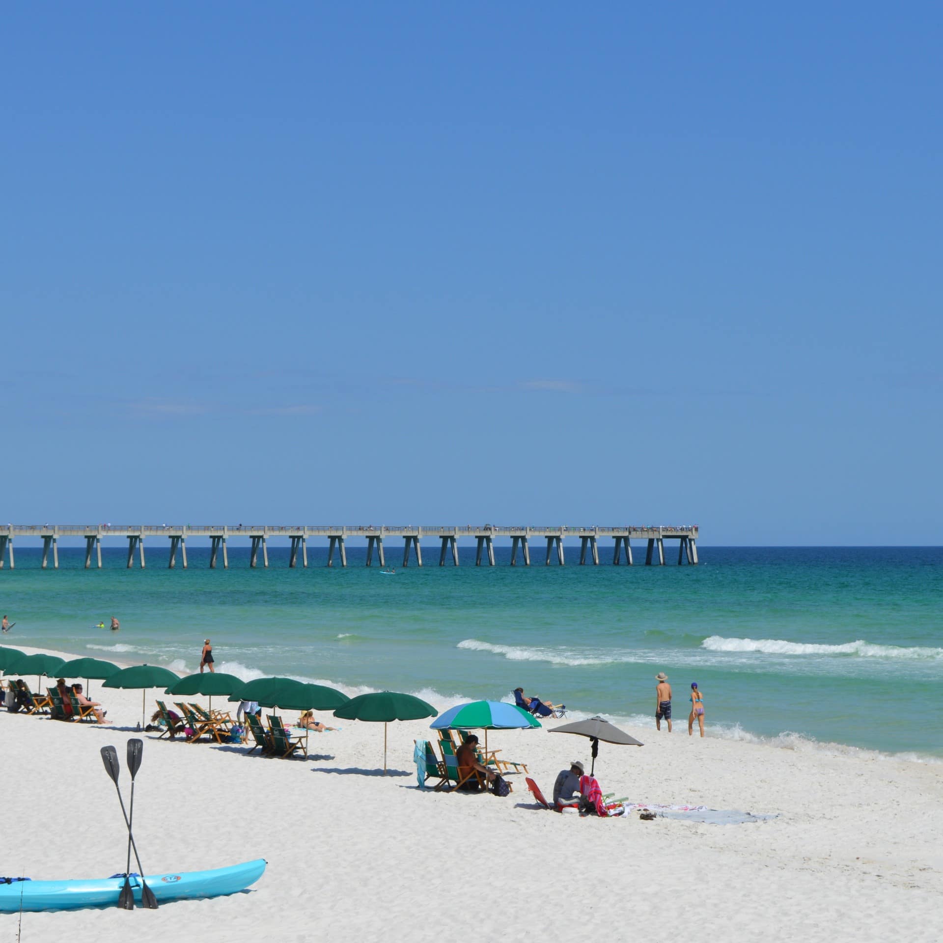 People walk up Fort Walton Beach's sands by kayaks, sun umbrellas, and a long fishing pier
